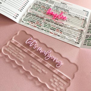 May include: Clear acrylic game pieces with the text "let's mahjong" in pink script. The image includes a game score sheet with numbers and text. The game pieces have a wavy edge and are designed for the game of Mahjong.