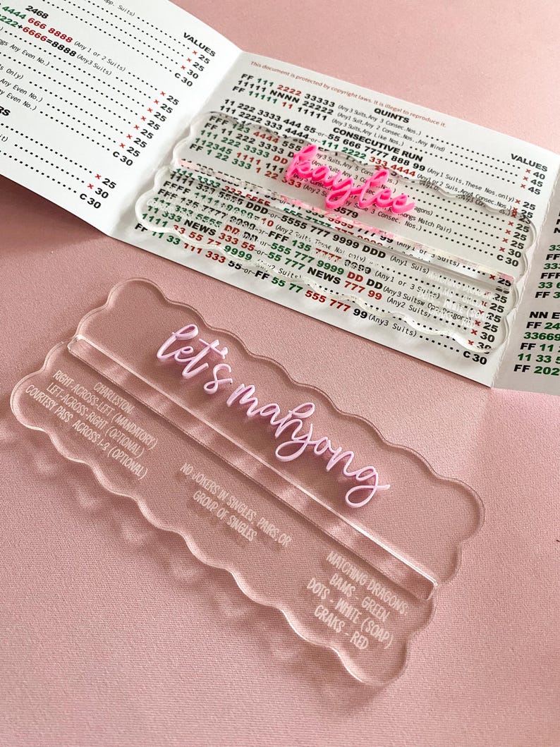 May include: Clear acrylic game pieces with the text "let's mahjong" in pink script. The image includes a game score sheet with numbers and text. The game pieces have a wavy edge and are designed for the game of Mahjong.