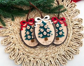 May include: Three wooden Christmas tree ornaments with family names and the year 2025. Each ornament features a green tree with white baubles, a brown trunk, and a red or white bow. The ornaments are on a woven mat.