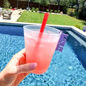 K&ouml;nnte beinhalten: Ein klarer Plastikbecher gef&uuml;llt mit einem rosa Getr&auml;nk und Eis, mit einem roten Strohhalm. Der Becher hat einen lila Halter mit der Aufschrift "KAYLEE". Das Getr&auml;nk wird in der N&auml;he eines Swimmingpools gehalten.