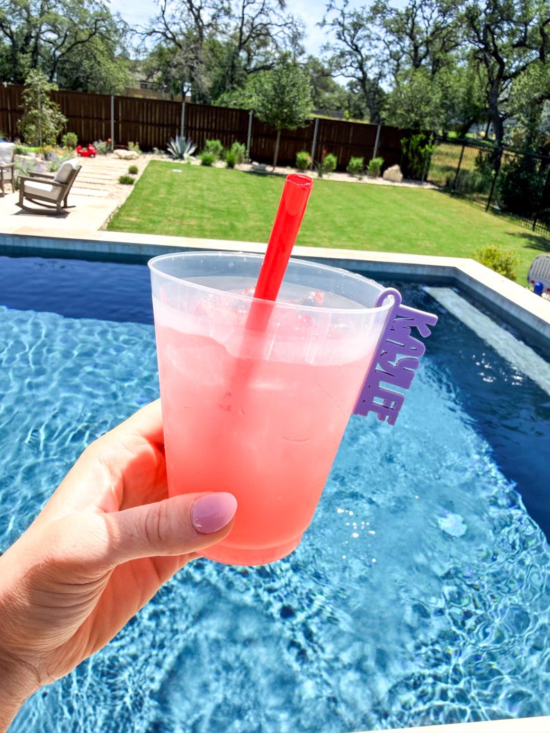 K&ouml;nnte beinhalten: Ein klarer Plastikbecher gef&uuml;llt mit einem rosa Getr&auml;nk und Eis, mit einem roten Strohhalm. Der Becher hat einen lila Halter mit der Aufschrift "KAYLEE". Das Getr&auml;nk wird in der N&auml;he eines Swimmingpools gehalten.