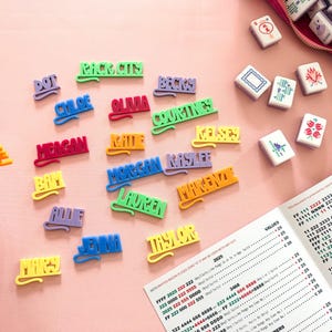 K&ouml;nnte beinhalten: Bunte Namensschilder und Mahjong-Steine ​​liegen auf einer rosa Oberfl&auml;che. Die Namensschilder in verschiedenen Farben schreiben Namen wie Chloe und Taylor. Ein Mahjong-Spiel-Scoreblatt ist ebenfalls sichtbar.