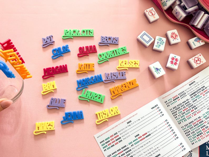 K&ouml;nnte beinhalten: Bunte Namensschilder und Mahjong-Steine ​​liegen auf einer rosa Oberfl&auml;che. Die Namensschilder in verschiedenen Farben schreiben Namen wie Chloe und Taylor. Ein Mahjong-Spiel-Scoreblatt ist ebenfalls sichtbar.