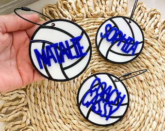 May include: Three white volleyball-shaped ornaments with black stitching and blue lettering. The ornaments read "Natalie", "Sophia", and "Coach Cassy".