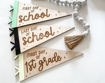 以下が含まれることがあります： ブラウンのフォントで「First Day of School」、「Last Day of School」、「First Day of 1st Grade」と書かれた木製のペナントフラッグ3つ。フラッグは小さな白い星で飾られており、緑、黒、黄色の縁取りがされています。