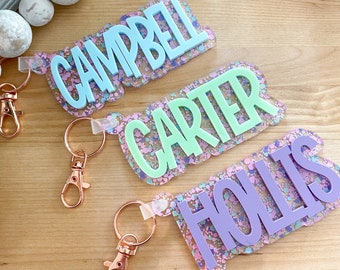 May include: Three personalized keychains with names "Campbell", "Carter", and "Hollis" written in white letters on a light blue, green, and purple background with glitter. Each keychain has a rose gold key ring.
