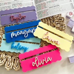 May include: A selection of brightly coloured acrylic name card holders in pink, blue, yellow, and purple, each with a name in a contrasting colour. Mahjong tiles and game score sheets are in the background.