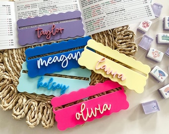 May include: A collection of colorful acrylic name card holders in pink, blue, yellow, and purple, each with a name in a contrasting color. Mahjong tiles and game score sheets are in the background.