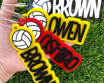 May include: A collection of colorful volleyball-themed name tags. The tags feature a volleyball graphic and names like "BROWN" and "OWEN" in bold, contrasting colors. Each tag is attached to a silver wire loop.