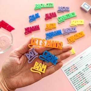 K&ouml;nnte beinhalten: Eine Sammlung bunter Namensschilder und Spielsteine aus Kunststoff auf einer rosa Oberfl&auml;che. Die Schilder tragen Namen wie "Chloe" und S&auml;tze wie "Let's Mahj". Spielsteine und eine gedruckte Anleitung sind ebenfalls zu sehen.