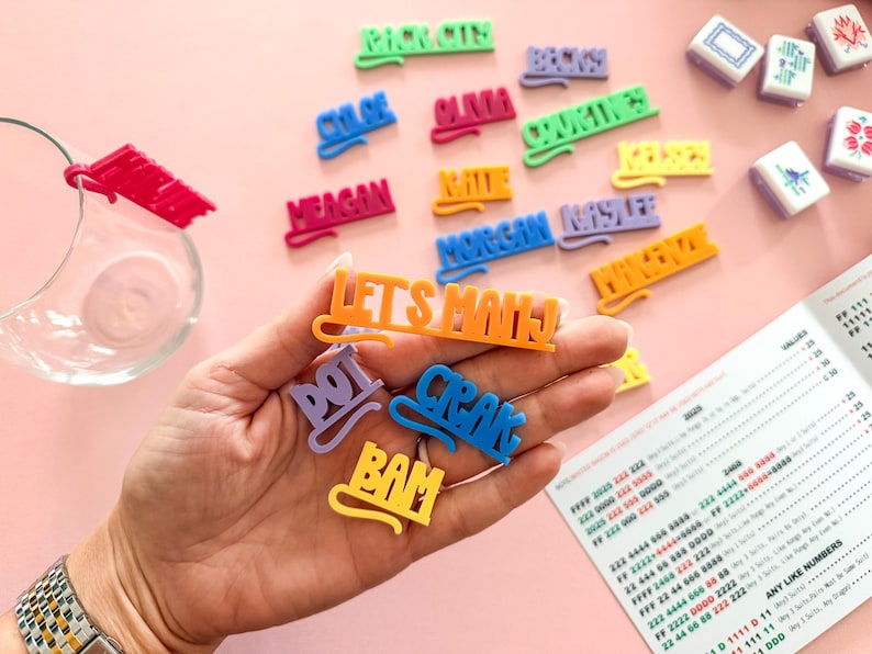 K&ouml;nnte beinhalten: Eine Sammlung bunter Namensschilder und Spielsteine aus Kunststoff auf einer rosa Oberfl&auml;che. Die Schilder tragen Namen wie "Chloe" und S&auml;tze wie "Let's Mahj". Spielsteine und eine gedruckte Anleitung sind ebenfalls zu sehen.