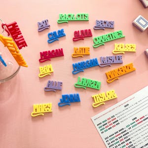K&ouml;nnte beinhalten: Bunte Namensschilder in verschiedenen Farben, darunter Rot, Gr&uuml;n und Gelb, liegen auf einer rosa Oberfl&auml;che. Die Namen sind in einer verspielten Schriftart gehalten. Eine Glasschale und ein Dokument mit Text sind ebenfalls sichtbar.