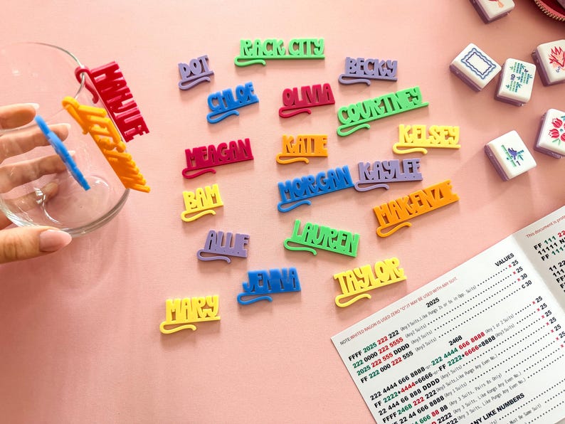 K&ouml;nnte beinhalten: Bunte Namensschilder in verschiedenen Farben, darunter Rot, Gr&uuml;n und Gelb, liegen auf einer rosa Oberfl&auml;che. Die Namen sind in einer verspielten Schriftart gehalten. Eine Glasschale und ein Dokument mit Text sind ebenfalls sichtbar.