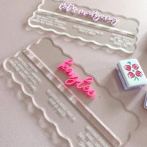 May include: Clear acrylic Mahjong scoreboards with pink lettering. One reads "let's mahjong" and the other "Kaylee". The boards have scalloped edges and include game instructions. A small tile with a floral design is also visible.