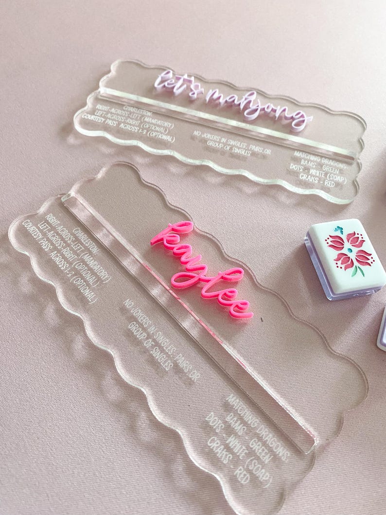 May include: Clear acrylic Mahjong scoreboards with pink lettering. One reads "let's mahjong" and the other "Kaylee". The boards have scalloped edges and include game instructions. A small tile with a floral design is also visible.