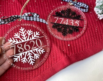 May include: Two clear round ornaments with a snowflake design and the text "There's no place like home for the holidays" and a house number. The ornaments are hanging from a red surface.