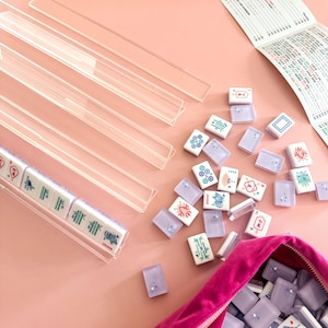 May include: A Mahjong set with white tiles featuring colorful illustrations and Chinese characters. The set includes clear acrylic tile racks, a pink velvet pouch, and a game guide, all arranged on a pink surface.