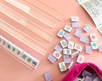 May include: A Mahjong set with white tiles featuring colorful illustrations and Chinese characters. The set includes clear acrylic tile racks, a pink velvet pouch, and a game guide, all arranged on a pink surface.