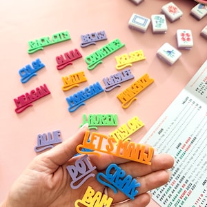 K&ouml;nnte beinhalten: Eine Sammlung farbenfroher Namensschilder und Spielsteine aus Kunststoff, darunter die Worte "LET'S MAHJ" und einzelne Namen. Die Tags sind in verschiedenen Farben gehalten, mit einer rosa Rei&szlig;verschlusstasche und Spielsteinen im Hintergrund. Ein Spielzettel ist ebenfalls sichtbar.