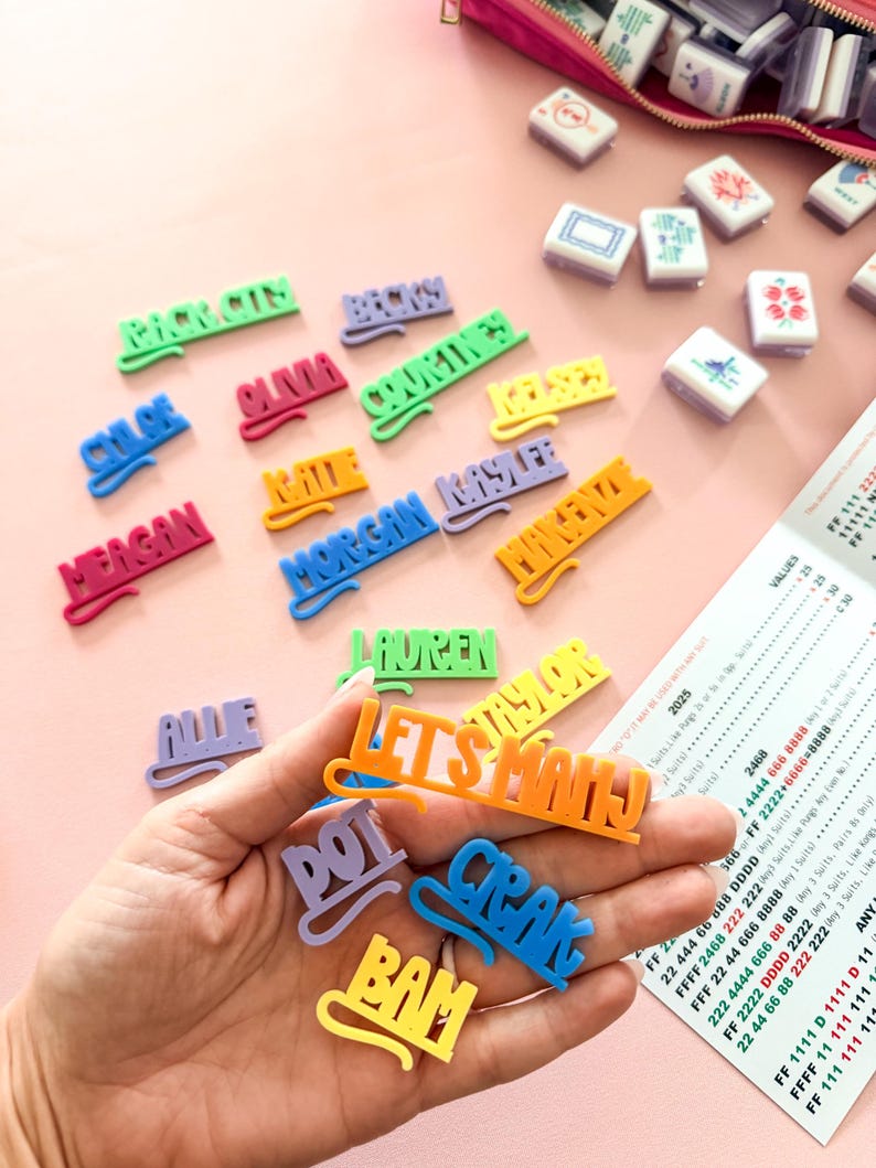 K&ouml;nnte beinhalten: Eine Sammlung farbenfroher Namensschilder und Spielsteine aus Kunststoff, darunter die Worte "LET'S MAHJ" und einzelne Namen. Die Tags sind in verschiedenen Farben gehalten, mit einer rosa Rei&szlig;verschlusstasche und Spielsteinen im Hintergrund. Ein Spielzettel ist ebenfalls sichtbar.