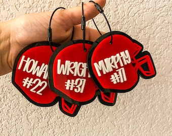 May include: Three red football helmet-shaped tags with black trim and black cords. Each tag features a white name and number: "HOWA #22", "WRIGHT #31", and "MURPHY #11". The tags are likely for sports fans or team members.