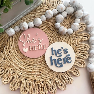 Puede incluir: Dos letreros redondos de madera con las frases "She's Here" y "He's Here" en letras blancas y azules. Los letreros están sobre un salvamanteles tejido, con una guirnalda de cuentas y una borla decorativa.