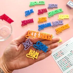 K&ouml;nnte beinhalten: Eine Sammlung farbenfroher, 3D-gedruckter Namensschilder und Phrasen, darunter "LET'S MAHJ", "DOT" und "CRAK". Die Tags sind in verschiedenen Farben wie Lila, Blau und Orange gehalten. Der Hintergrund ist eine hellrosa Oberfl&auml;che.