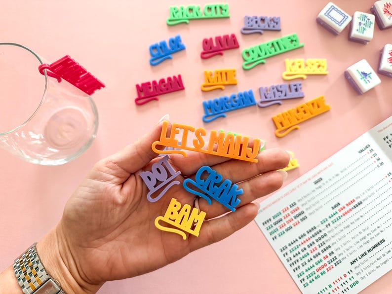 K&ouml;nnte beinhalten: Eine Sammlung farbenfroher, 3D-gedruckter Namensschilder und Phrasen, darunter "LET'S MAHJ", "DOT" und "CRAK". Die Tags sind in verschiedenen Farben wie Lila, Blau und Orange gehalten. Der Hintergrund ist eine hellrosa Oberfl&auml;che.