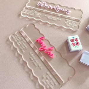 May include: Clear acrylic Mahjong tile holders with pink text that reads "let's mahjong" and "Kaylee". The holders have a scalloped edge and include game instructions. A Mahjong tile with a floral design is visible.
