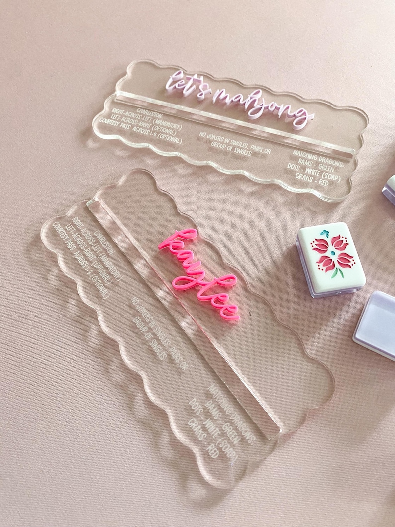 May include: Clear acrylic Mahjong tile holders with pink text that reads "let's mahjong" and "Kaylee". The holders have a scalloped edge and include game instructions. A Mahjong tile with a floral design is visible.