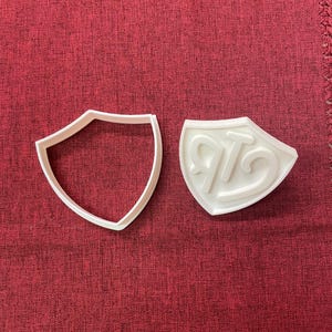 Könnte beinhalten: Zwei weiße Ausstecher in Schildform. Einer ist eine durchgezogene Umrisslinie, der andere hat die Buchstaben "RTG" geprägt. Die Ausstecher liegen auf einer roten, strukturierten Oberfläche.