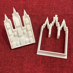 Könnte beinhalten: Zwei weiße Ausstecher aus Kunststoff in Form eines Schlosses. Einer ist eine Vollform mit Innendetails, der andere eine Kontur. Die Ausstecher liegen auf einer roten, strukturierten Oberfläche.