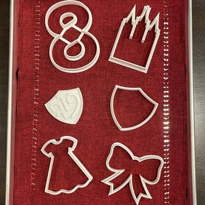 Könnte beinhalten: Weiße Ausstecher aus Kunststoff in verschiedenen Formen, darunter die Zahl Acht, ein Schloss, zwei Schilde, ein Kleid und eine Schleife. Die Ausstecher sind auf einem roten Stoff in einer Metallbackform angeordnet.