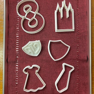 Könnte beinhalten: Ein Set mit sieben weißen Plastik-Ausstechformen auf einer roten Stoffoberfläche. Die Ausstechformen sind in Form einer Acht, einer Kirche, eines Schildes, eines Kleides, einer Krawatte und zweier verschiedener Schilde.