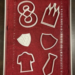 Könnte beinhalten: Eine Sammlung weißer Ausstecher aus Kunststoff in verschiedenen Formen, darunter die Zahl Acht, eine Krone, zwei Schilde, ein T-Shirt und eine Krawatte. Die Ausstecher sind auf einem rot stoffausgekleideten Metalltablett angeordnet.