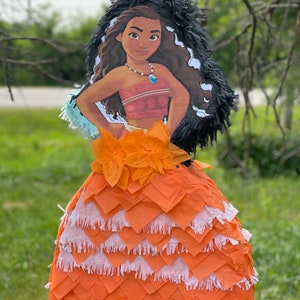 Pu&ograve; includere: Una pignatta a forma di personaggio dei cartoni animati, raffigurante una donna dalla pelle scura con lunghi capelli castani ondulati. La pignatta ha una gonna a strati arancione e bianca, un top rosso e un copricapo a frange nere. &Egrave; appesa a un ramo.
