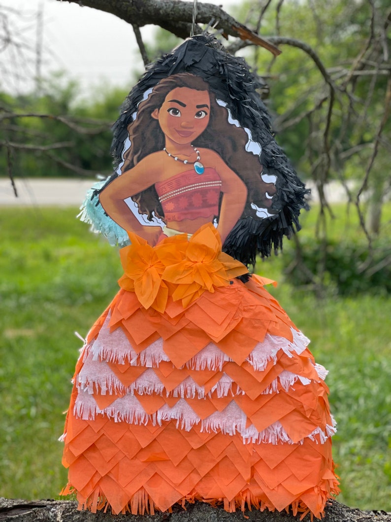 Pu&ograve; includere: Una pignatta a forma di personaggio dei cartoni animati, raffigurante una donna dalla pelle scura con lunghi capelli castani ondulati. La pignatta ha una gonna a strati arancione e bianca, un top rosso e un copricapo a frange nere. &Egrave; appesa a un ramo.