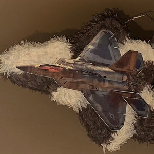 May include: A detailed paper model of an F-22 Raptor fighter jet, featuring camouflage patterns in shades of gray, brown, and blue. The jet is mounted on a decorative base with white and brown fringed textures, creating a layered effect.