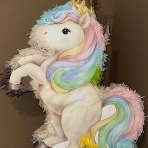 May include: A whimsical unicorn piñata with a white body, a golden horn, and a colorful mane and tail. The mane and tail feature pastel shades of blue, pink, yellow, and green. The piñata is decorated with fringed paper and has a smiling expression.