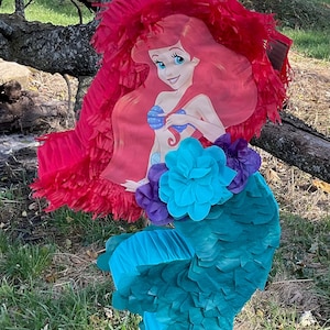 May include: Ariel-themed piñata featuring a red fringe top, a turquoise tail, and a central image of Ariel. The piñata is decorated with purple and turquoise paper flowers and is designed for a party or celebration.