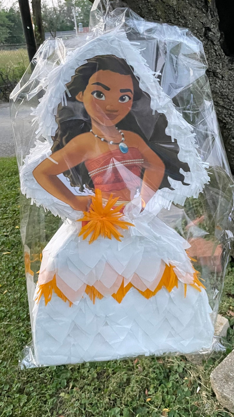 Pu&ograve; includere: Una pignatta a tema Moana, con un'illustrazione a cartoni animati del personaggio in un vestito bianco e arancione. La pignatta &egrave; avvolta in plastica trasparente, con una frangia bianca intorno alla testa.