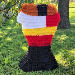 May include: A piñata featuring horizontal layers of fringed paper in orange, black, white, yellow, and red. The structure has a wide top section that tapers inward toward the middle before widening again at the base, creating a distinct hourglass shape.