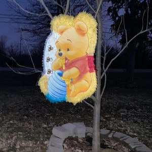 May include: A colorful Winnie the Pooh decoration, featuring the beloved bear in a red shirt, eating honey from a blue pot. The decoration is hanging from a tree branch, with a night sky background.