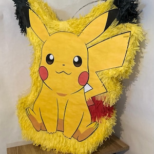 May include: A yellow Pikachu piñata with black-tipped ears, red cheeks, and a smiling face. The piñata is made of yellow crepe paper and has a red and yellow tail. It is designed to look like the popular Pokémon character.