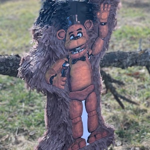 May include: A brown Freddy Fazbear piñata, featuring the character with a black top hat, bow tie, and microphone. The piñata is made of brown paper fringe and is hanging outdoors.