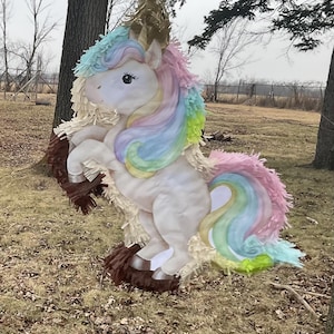 May include: A unicorn piñata featuring a white body with a pastel rainbow mane and tail. The design includes fringed paper accents in shades of cream, brown, and light blue. The unicorn is depicted in a rearing pose with a gold horn on its head.