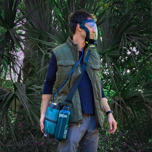 May include: A black and clear respirator mask with a blue strap, connected to a teal-colored portable air supply unit. The unit includes a water bottle with the text "RDA". The person is wearing a green vest and a blue shirt.