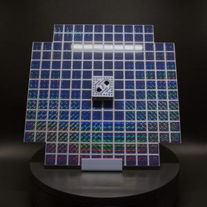 May include: A square, mosaic-like art piece with a grid of small, iridescent blue squares. A small, white square with a circuit board design is centered on the piece. The artwork is mounted on a black, circular base.