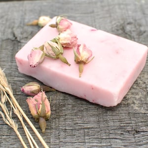 Peut inclure: Un pain de savon rectangulaire rose orné de boutons de roses séchées. Le savon est posé sur une surface en bois vieilli, avec des herbes séchées à gauche. Le savon semble fait à la main et a une esthétique naturelle.