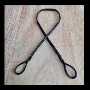 Puede incluir: Una cuerda trenzada negra con dos bucles en los extremos. La cuerda está tendida sobre una superficie de madera.
